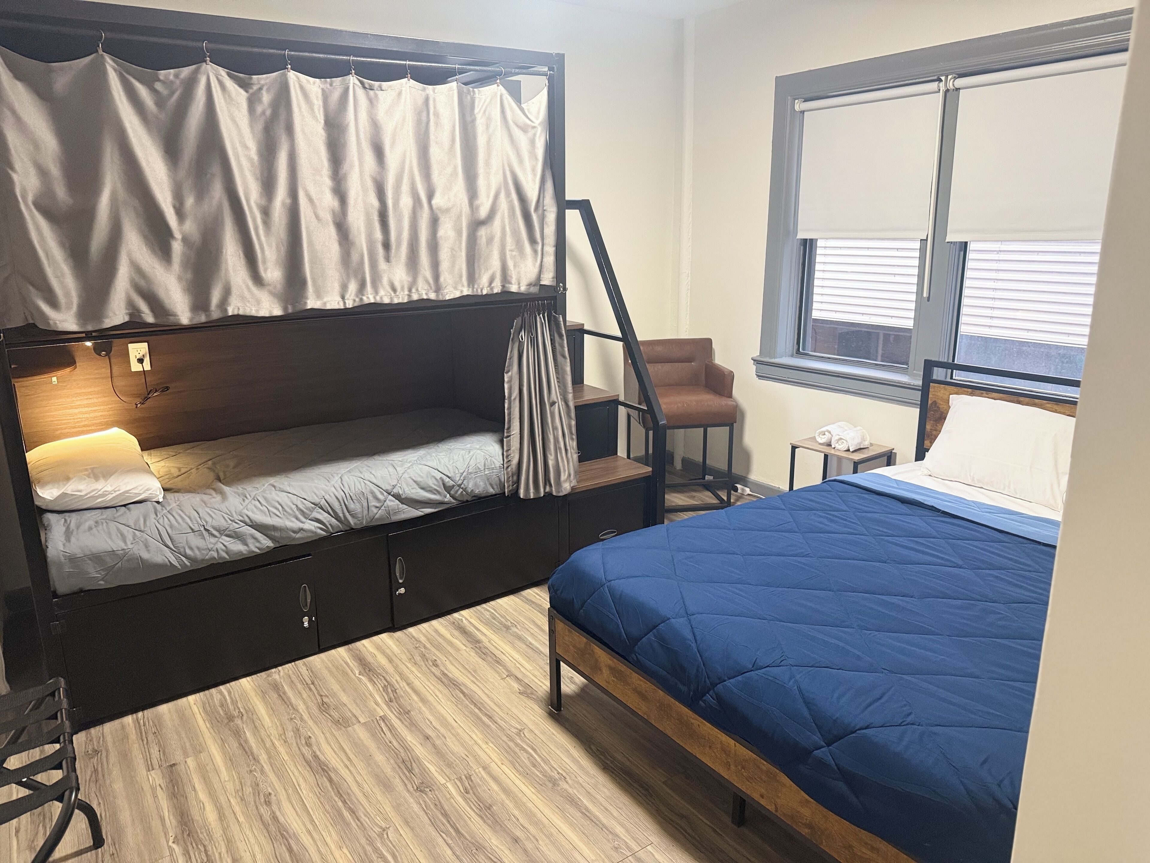 Habitación compacta con literas, una cama individual, una silla, una mesita y una ventana con persianas.