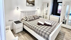 Un dormitorio moderno con una cama grande, mesitas de noche blancas, una mesita auxiliar, una ventana con cortinas y una lámpara fijada a la pared.