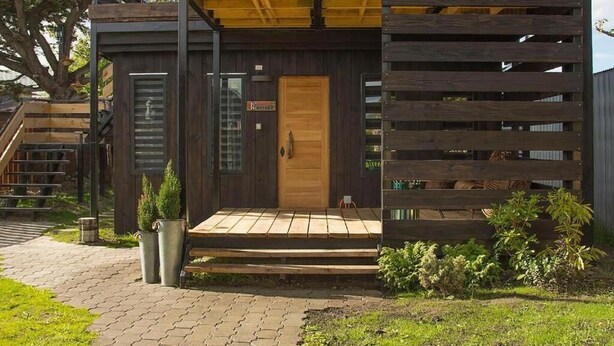 Eine moderne Holzhütte mit Holztür, einer kleinen Veranda und einer Metallbank.