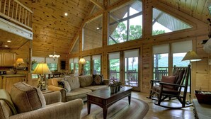 A spacious wooden cabin interior with a high ceiling, large windows, a cozy seating area, and a rocking chair.