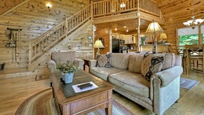 A wooden cabin interior with a living room, staircase, and kitchen area.