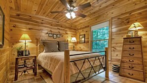 A wooden cabin bedroom with a bed, two bedside lamps, a dresser, and a window with a view of trees.