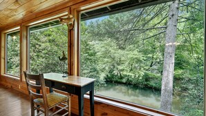 A wooden cabin room with large windows overlooking a forested area and a river.