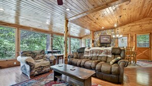 A cozy cabin interior with wooden ceilings, large windows, a stone fireplace, and rustic furnishings.