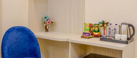 A compact room with a blue chair, a small round table with fruit, a refrigerator, a shelf with snacks, and a vase with flowers.