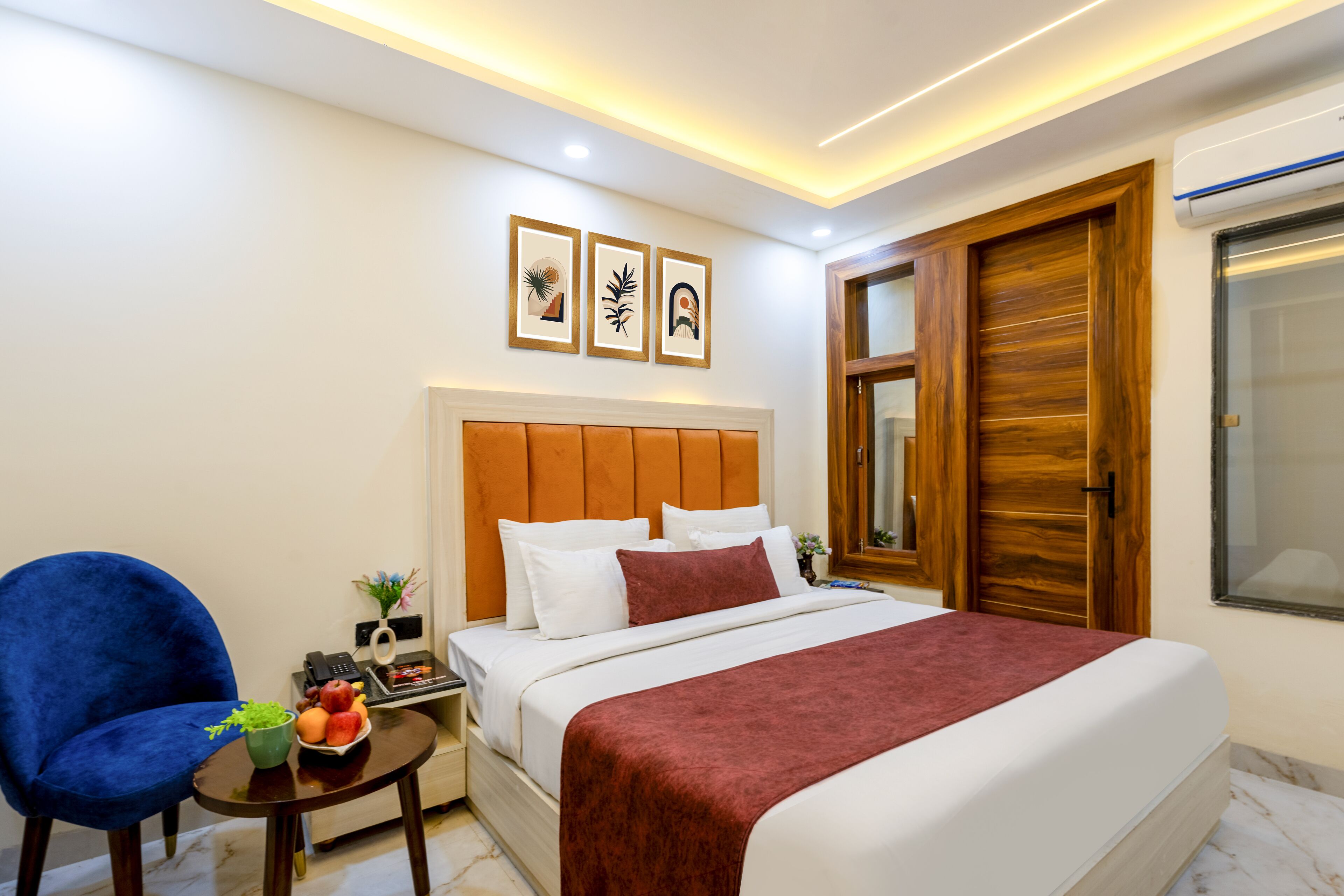 Une chambre d’hôtel avec un grand lit, une chaise bleue, une petite table sur laquelle se trouvent un panier de fruits et un vase, ainsi qu’une armoire en bois.