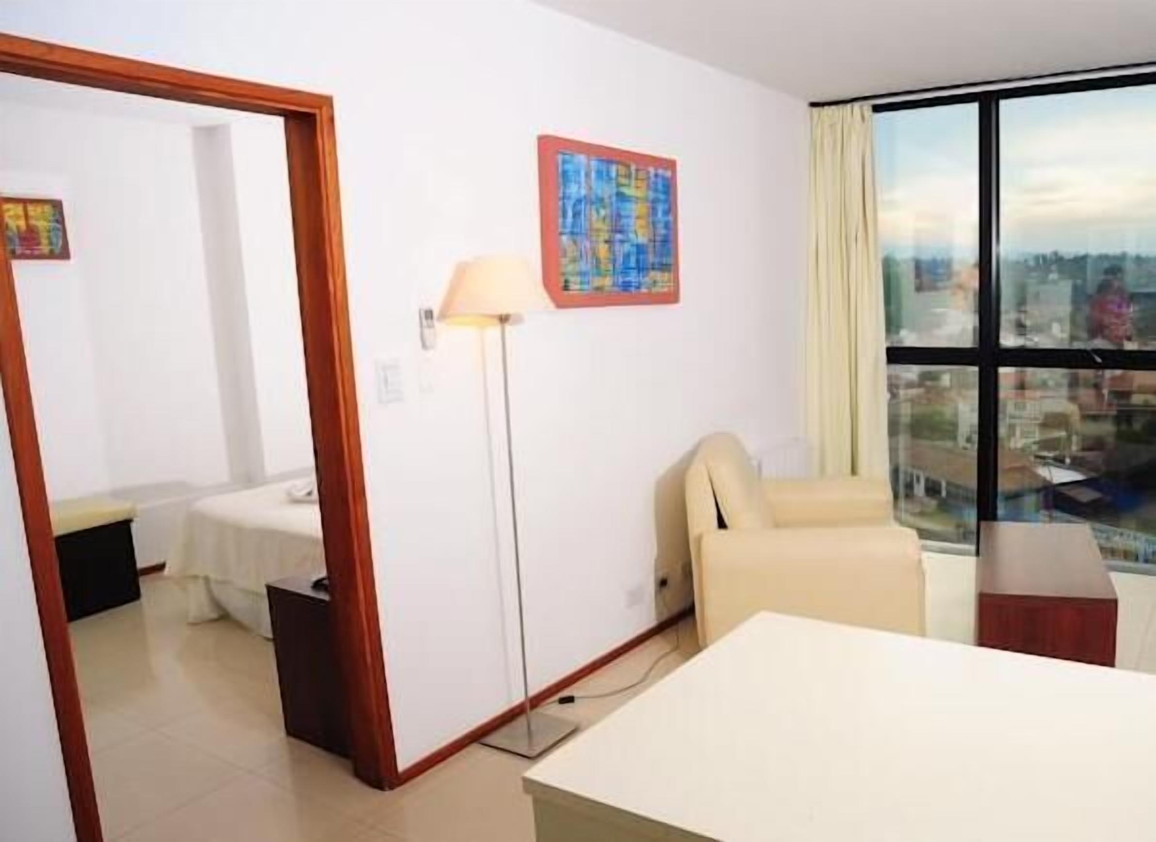 Une chambre d’hôtel avec un lit, une chaise, un bureau, une lampe et une fenêtre donnant sur la ville.