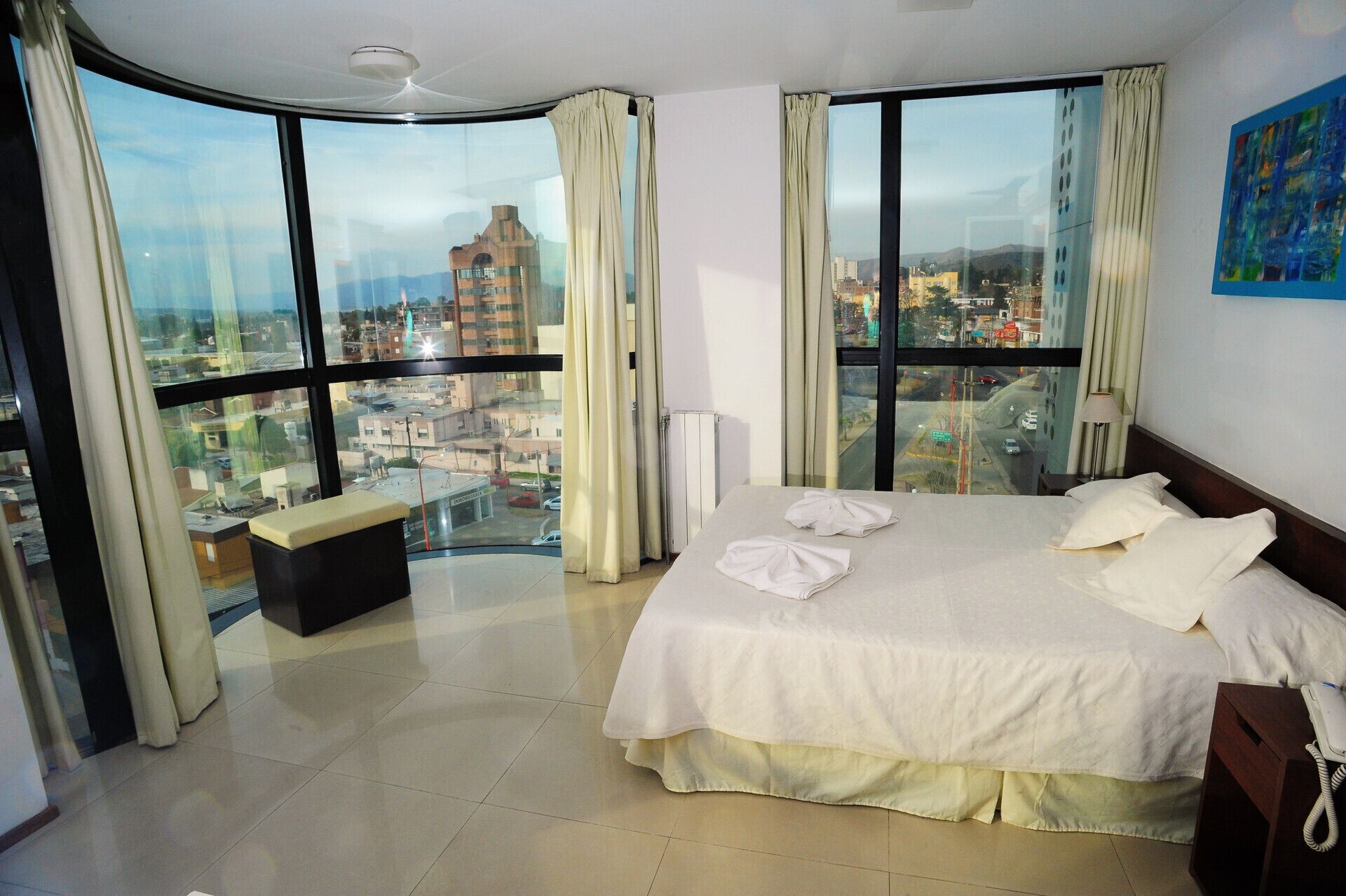 Habitación de hotel con una cama grande, una mesita de noche, una silla y vistas a la ciudad a través de amplias ventanas.