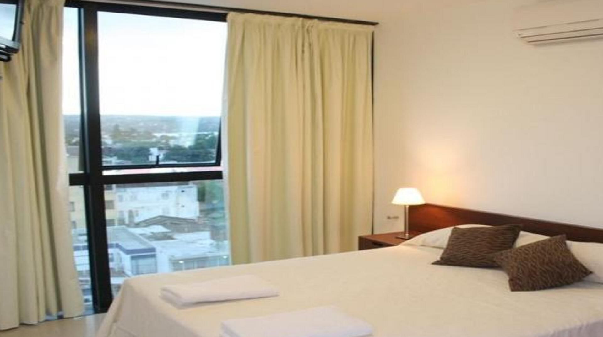 Habitación de hotel con un ventanal grande, cama con almohadas, mesita de noche con lámpara y vista a edificios.