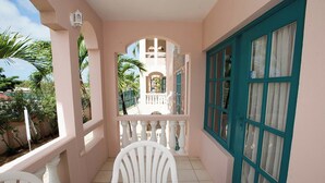 A balcony with a white table and chairs, a teal-framed window, and a view of a building with arches. - Charming 2Br, 2 bath Resort Condo,AC,pools,hot tubs,1M to main strip/beach,Aruba (Aruba)