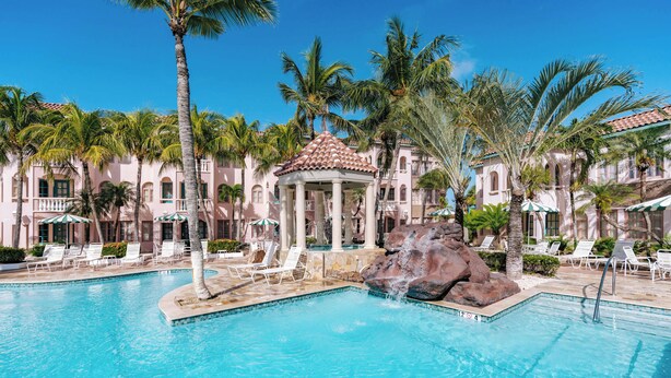 A pool area with lounge chairs, a gazebo, palm trees, and a building with a tiled roof. - Charming 2Br, 2 bath Resort Condo,AC,pools,hot tubs,1M to main strip/beach,Aruba (Aruba)