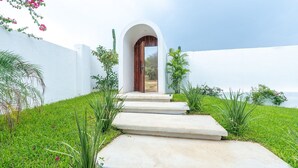 Un sentier mène à une porte en bois encadrée d’une arche blanche, le tout étant entour é de verdure et de cactus.
