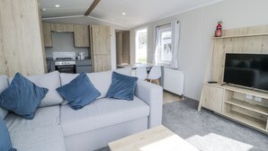 A modern living room with a grey sofa, blue cushions, a wooden coffee table, and a TV on a wooden stand. - Manor House Gold 2 Bedroom (Maryport)
