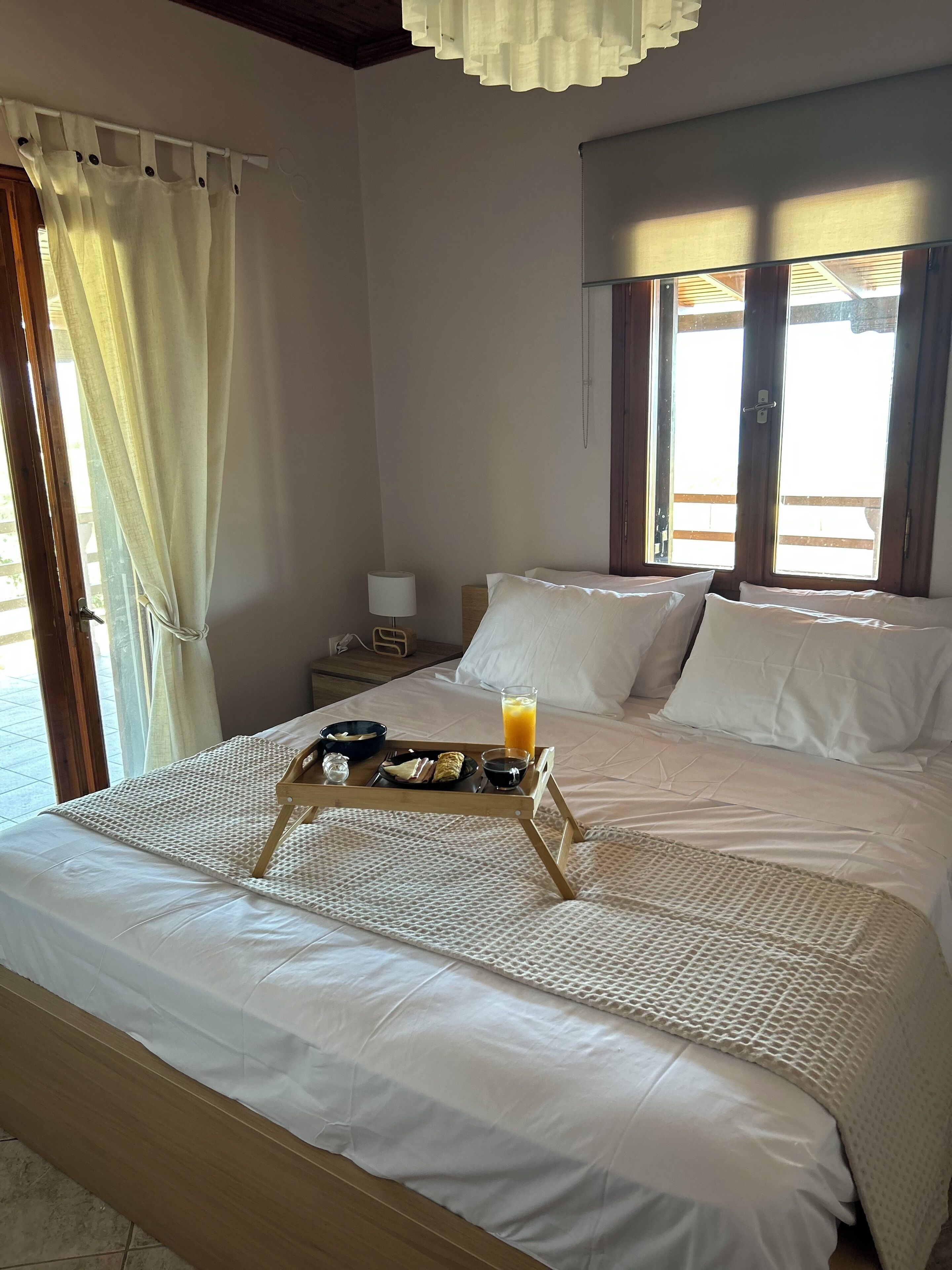 A neatly made bed with a tray of breakfast items, including a glass of orange juice, in a room with large windows and a view of the outdoors.