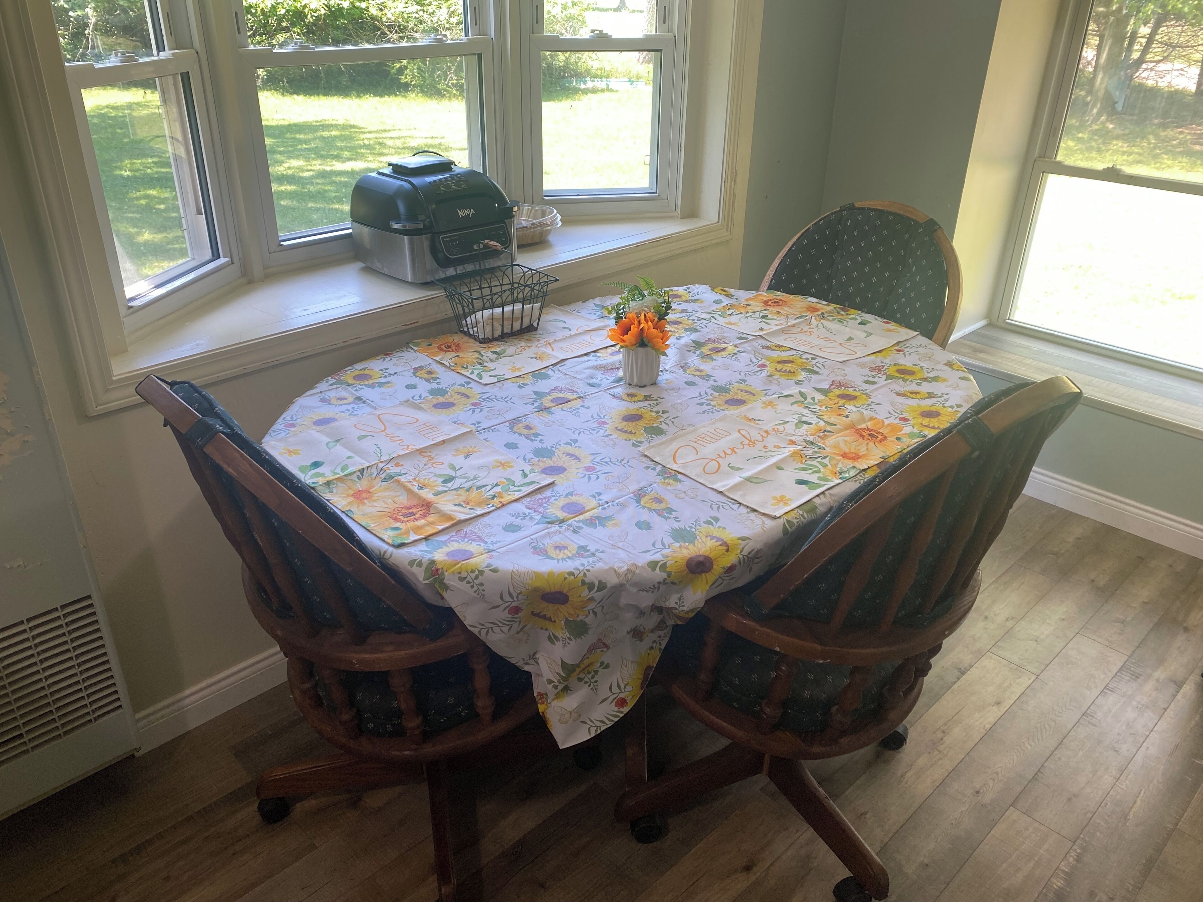 Une table à manger recouverte d’une nappe à motifs floraux, deux chaises en bois, une fenêtre avec des barreaux et un petit vase avec des fleurs.