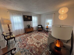 Un salon avec un tapis à motifs, un téléviseur à écran plat, un canapé, un fauteuil, une petite table avec une lampe et une plante décorative.