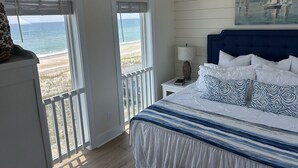 A bedroom with a bed, a nightstand, a lamp, a painting, and a view of the beach.