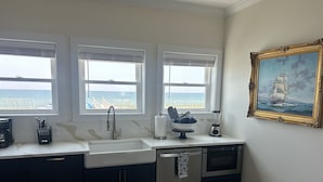 A kitchen with three windows overlooking the ocean, a coffee maker, a blender, and a painting on the wall.