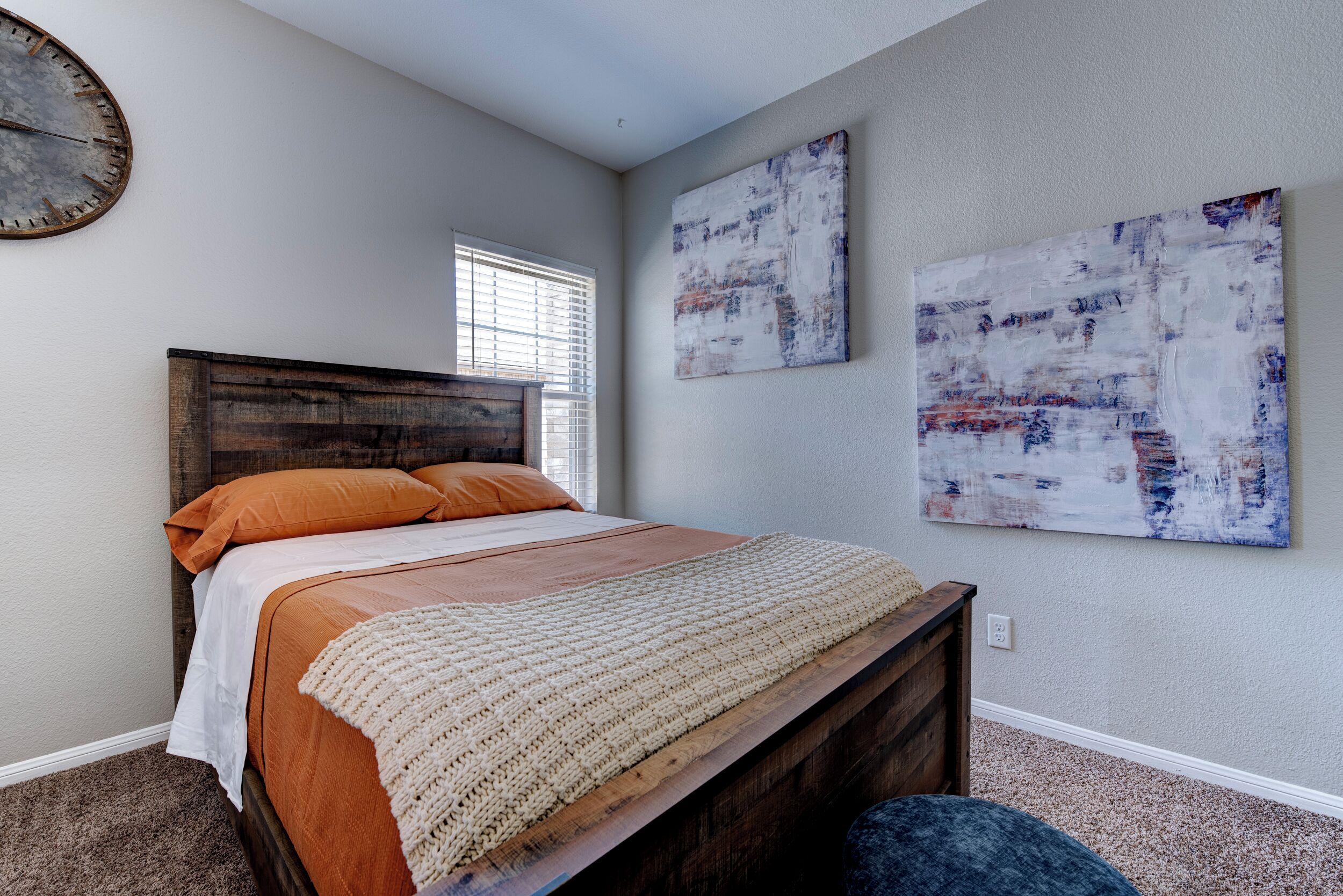 Une chambre avec un lit en bois, une literie orange, un plaid beige et des œuvres d’art abstraites accrochées au mur.