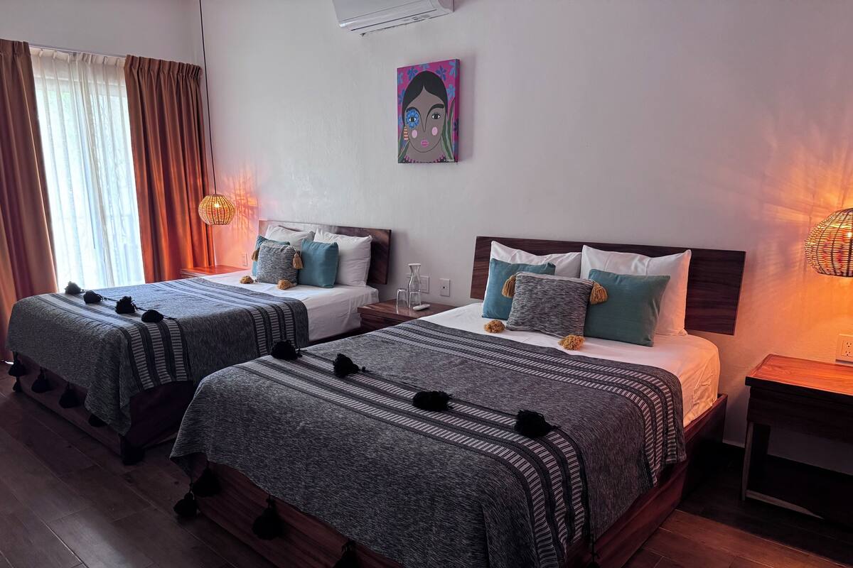 Habitación de hotel con dos camas, cada una con colcha y almohadas a rayas grises a juego. Hay un cuadro en la pared sobre las camas y una mesita de noche con lámpara al lado de la cama derecha.