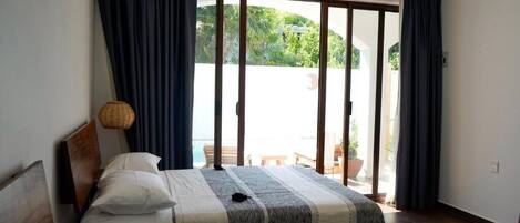 A room with two beds, a large sliding glass door leading to an outdoor area, and a wicker lamp on a bedside table.