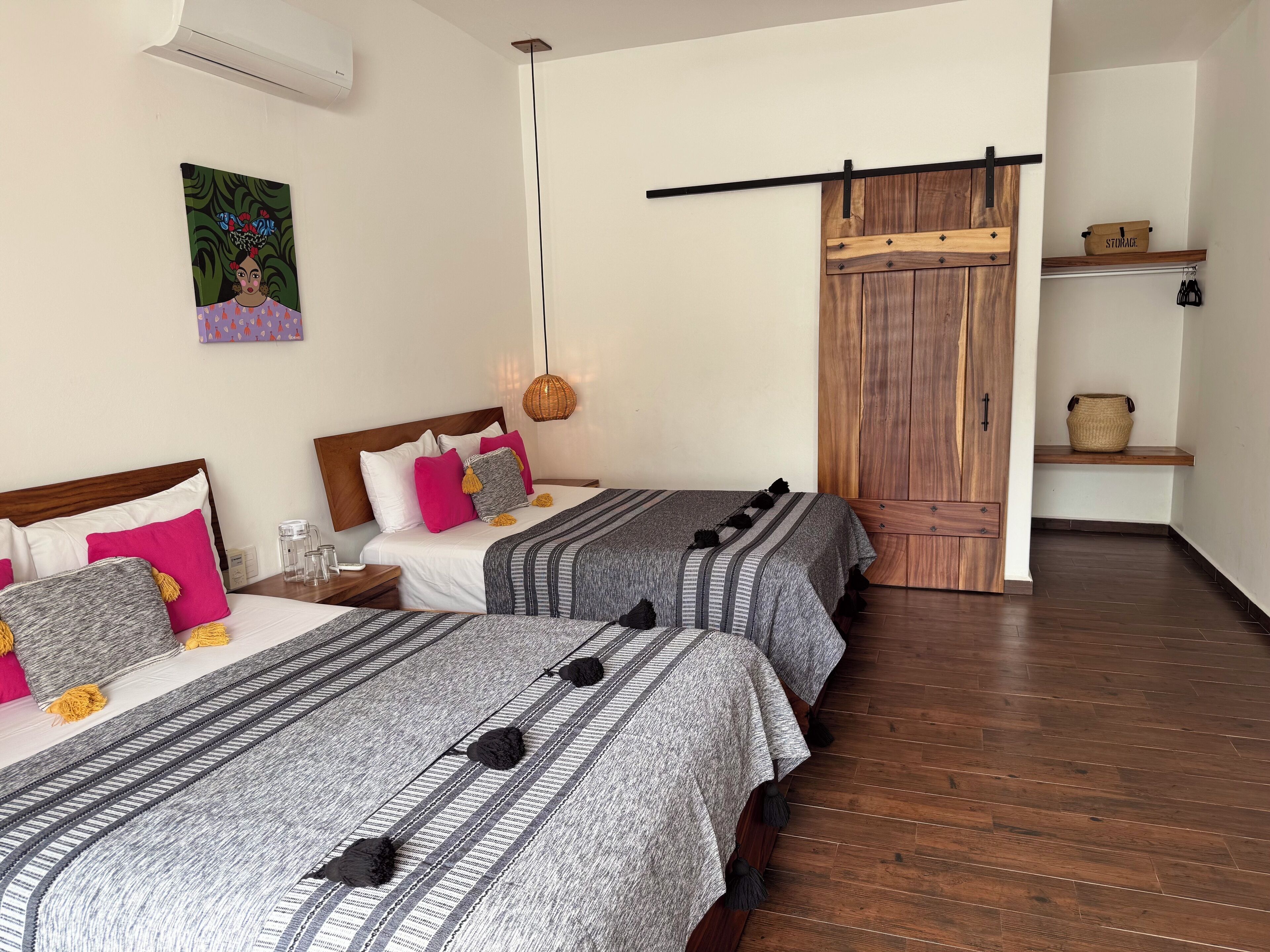 A hotel room with two beds, a wooden wardrobe, a hanging light fixture, and a small shelf with a basket.