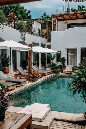 A pool area with lounge chairs and umbrellas, surrounded by potted plants and a white building. - Casa Beu - Adults Only (Santa Maria Colotepec)