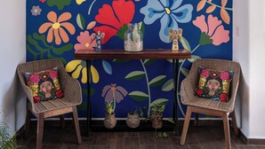 Quarto com um grande mural floral na parede, duas cadeiras de vime com almofadas coloridas, uma pequena mesa de madeira e plantas em vasos.