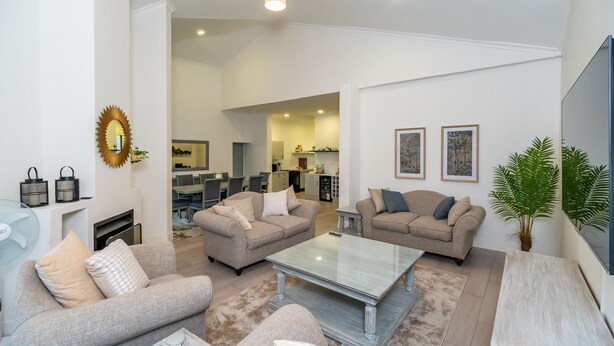 A spacious living room with two sofas, a coffee table, and a TV. There is a kitchen in the background and a dining area visible through an open doorway. - Stunning 3 Bedroom House Near Harare Airport -2297 (Harare)