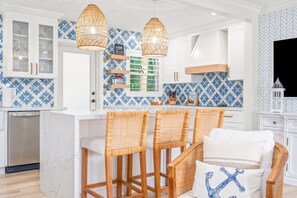A modern kitchen with white cabinets, blue patterned backsplash, wicker chairs, and pendant lights.