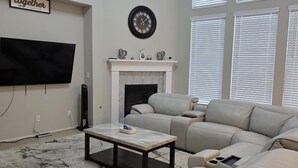 A living room with a beige sofa, a coffee table, a fireplace, a mounted TV, and a large window with blinds. - Family-Friendly 5BR Home, Game Room + Large Backyard! with AC, WiFi in 77407 (Richmond)