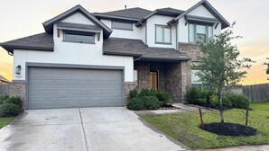 A two-story house with a grey garage door, a brick entrance, and a small front yard with a tree and shrubs. - Family-Friendly 5BR Home, Game Room + Large Backyard! with AC, WiFi in 77407 (Richmond)