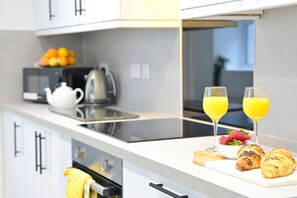A modern kitchen with a white countertop, a kettle, a toaster, and a cutting board with croissants and strawberries. There are two glasses of orange juice on the counter.