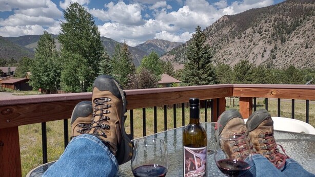 A person relaxing on a deck with a mountain view, holding a wine glass, and wearing hiking boots. - Three Eagle Nest - Mountains of Fun in Lake City, Colorado (Lake City)