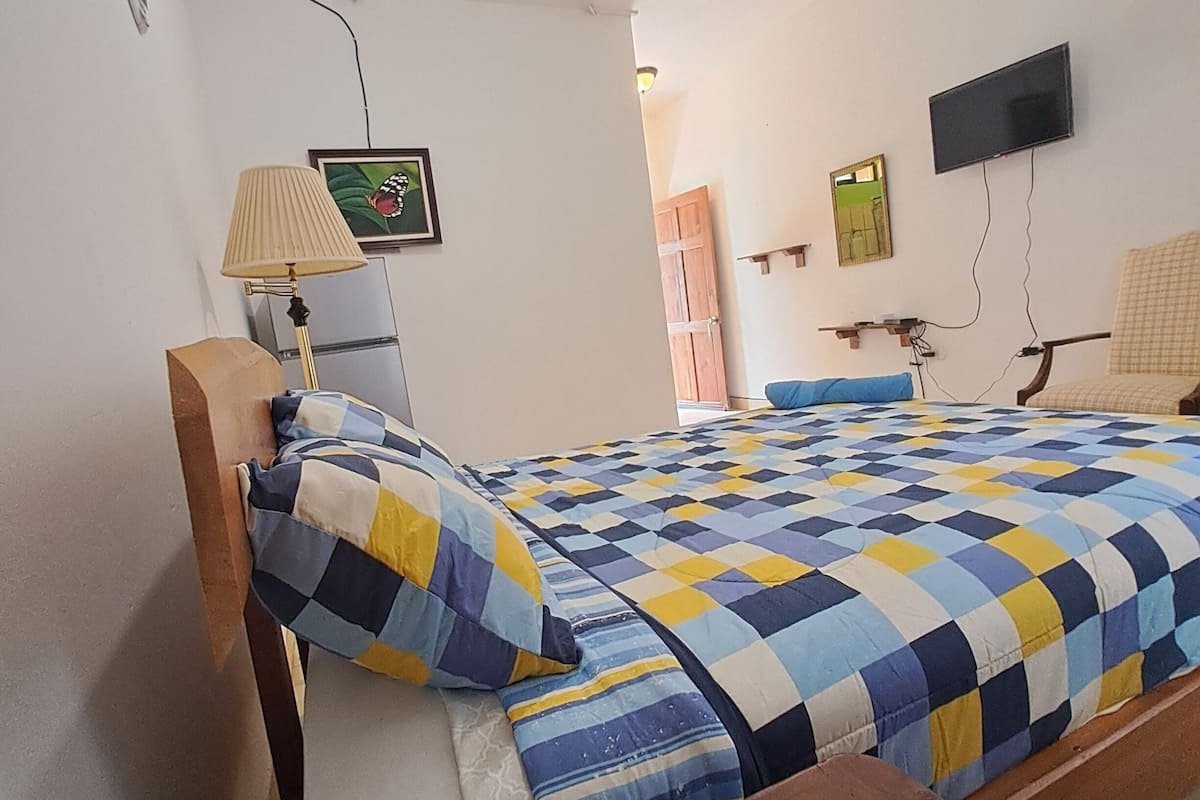 A bedroom with a bed, a bedside lamp, a TV mounted on the wall, and a door leading to another room.