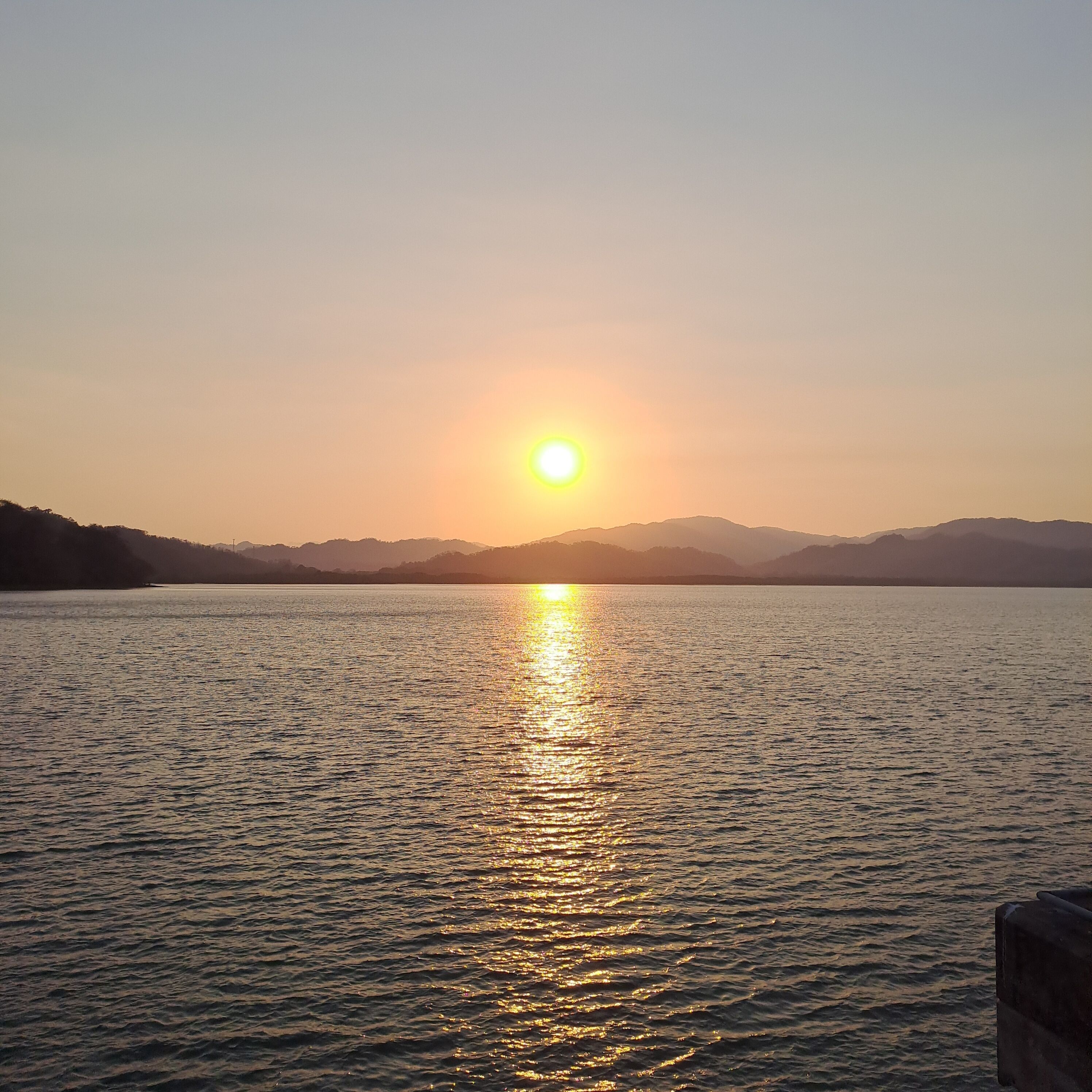 Um lago calmo com o sol refletindo na água, rodeado por colinas.