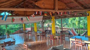 An outdoor seating area with wooden tables and chairs, a cushioned sofa, and a tiled floor. - Georgi, Junquillal (Veintisiete de Abril)