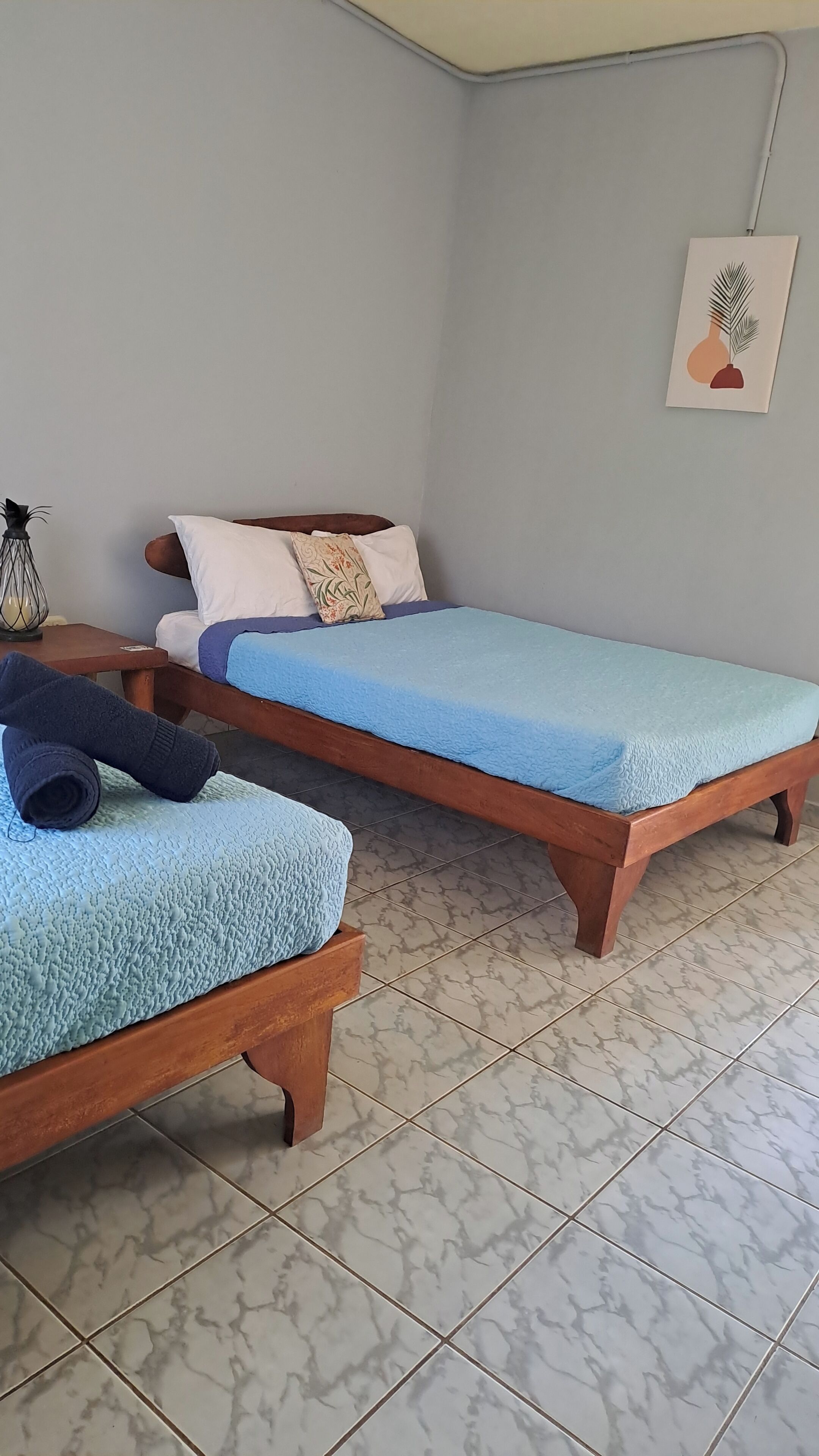 Dos camas de madera con ropa de cama azul, una mesita con una lámpara y una obra de arte colgada en la pared.