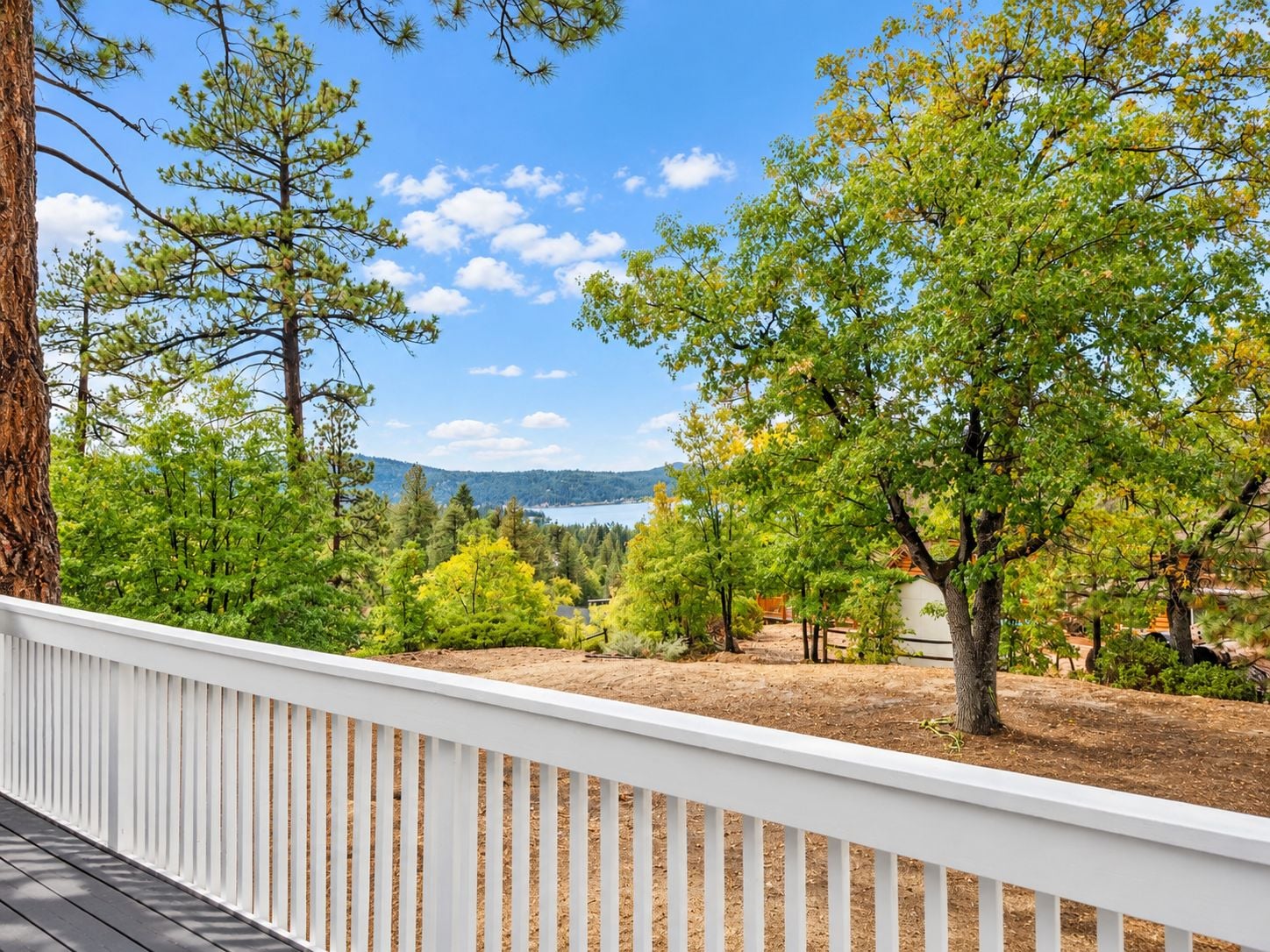 A deck with a white railing overlooking a landscaped area with trees and a view of a lake and mountains.