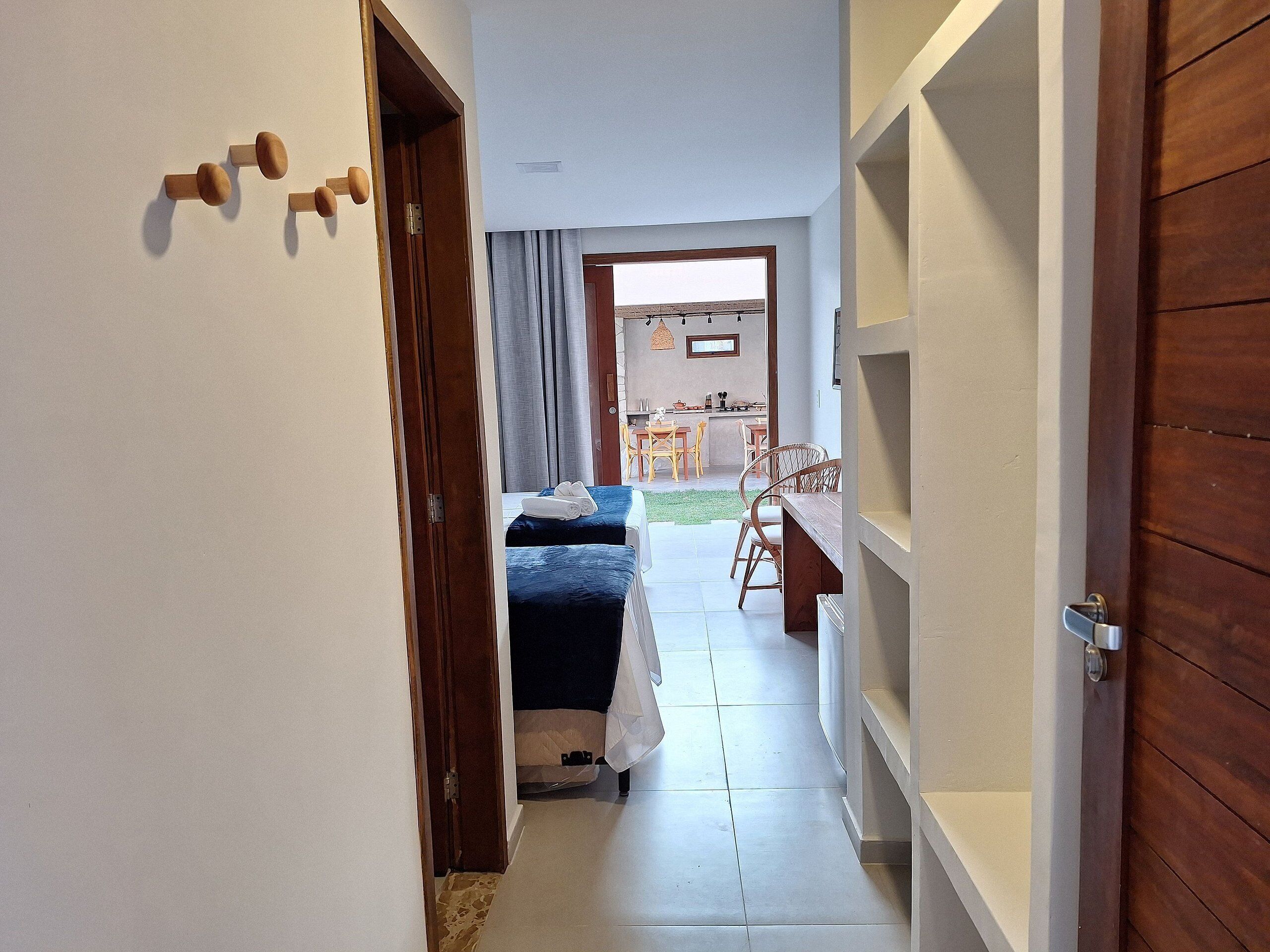 A narrow hallway leading to a bedroom with a bed, a small table, and a chair. There is a door on the left and a wooden door on the right.