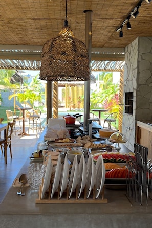 A buffet setup with various food items and utensils, a hanging woven light fixture, and a view of the outdoors. - Pousada Oasis dos Ventos (Touros)