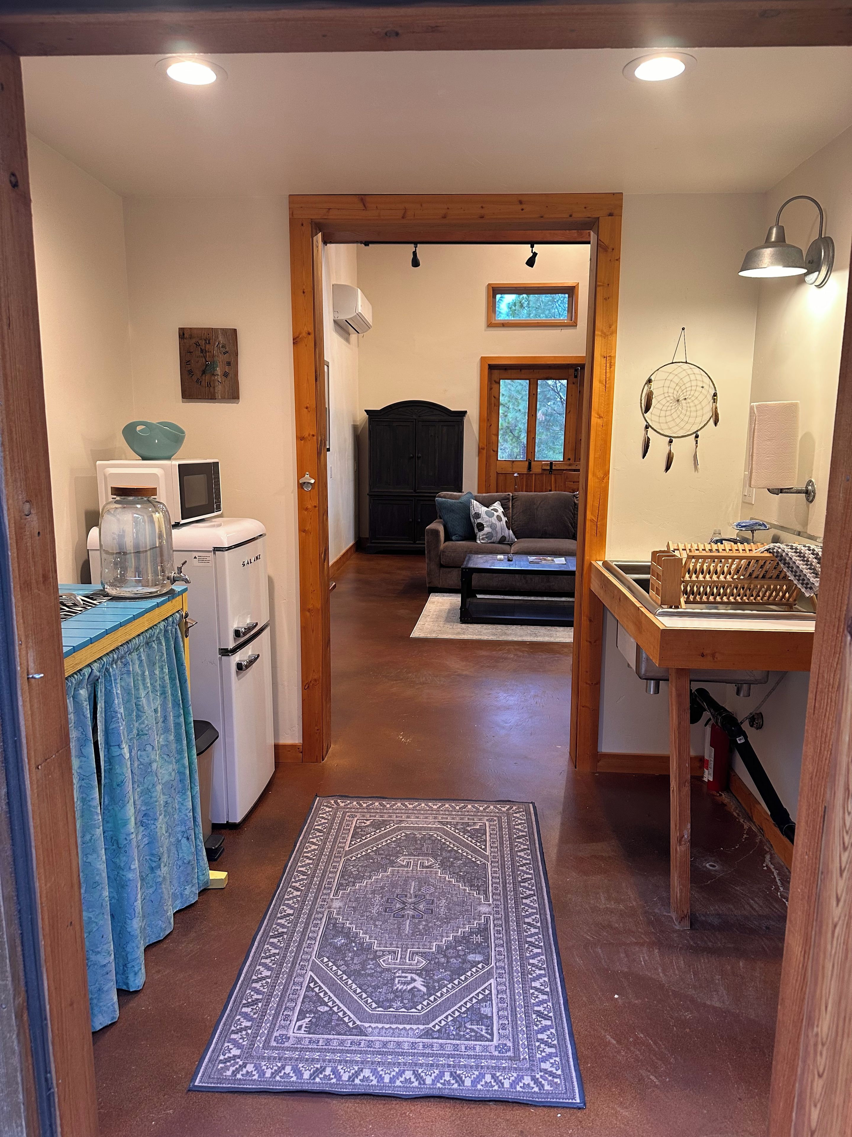 A hallway leading to a living area with a sofa, a wooden table, and a kitchen area with a refrigerator and microwave.