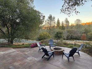 An outdoor patio with Adirondack chairs and a fire pit, surrounded by trees and a white fence.