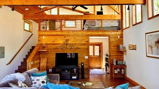 A spacious living room with a high ceiling and wooden beams, a staircase leading to an upper level, and a cozy seating area with a fireplace. - Rustic Studio Getaway in the California Gold Country - Pet Friendly (Penn Valley)