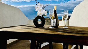 Una mesa con una botella de vino, dos copas y un objeto circular negro sobre una cubierta de madera, con vistas al cielo y edificios.