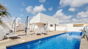 Una piscina con sillones de descanso y sombrillas, un edificio blanco y un cielo despejado.