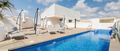 A swimming pool with lounge chairs and umbrellas, a white building, and a clear sky.