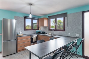 Une cuisine moderne équipée d’un réfrigérateur en acier inoxydable, d’un four encastré, d’un four à micro-ondes et d’un évier. On y trouve un grand îlot central avec une table et des chaises, et les murs sont peints dans une teinte turquoise, tandis que de grandes fenêtres illuminent la pièce.