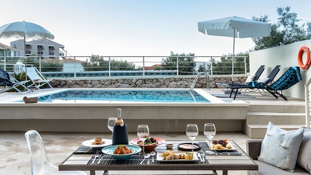 A poolside dining area with a table set for two, a pool, lounge chairs, and umbrellas. - Apartment 'Evagelia Luxury  3' with Shared Pool, Wi-Fi and Air Conditioning (?e??ada)
