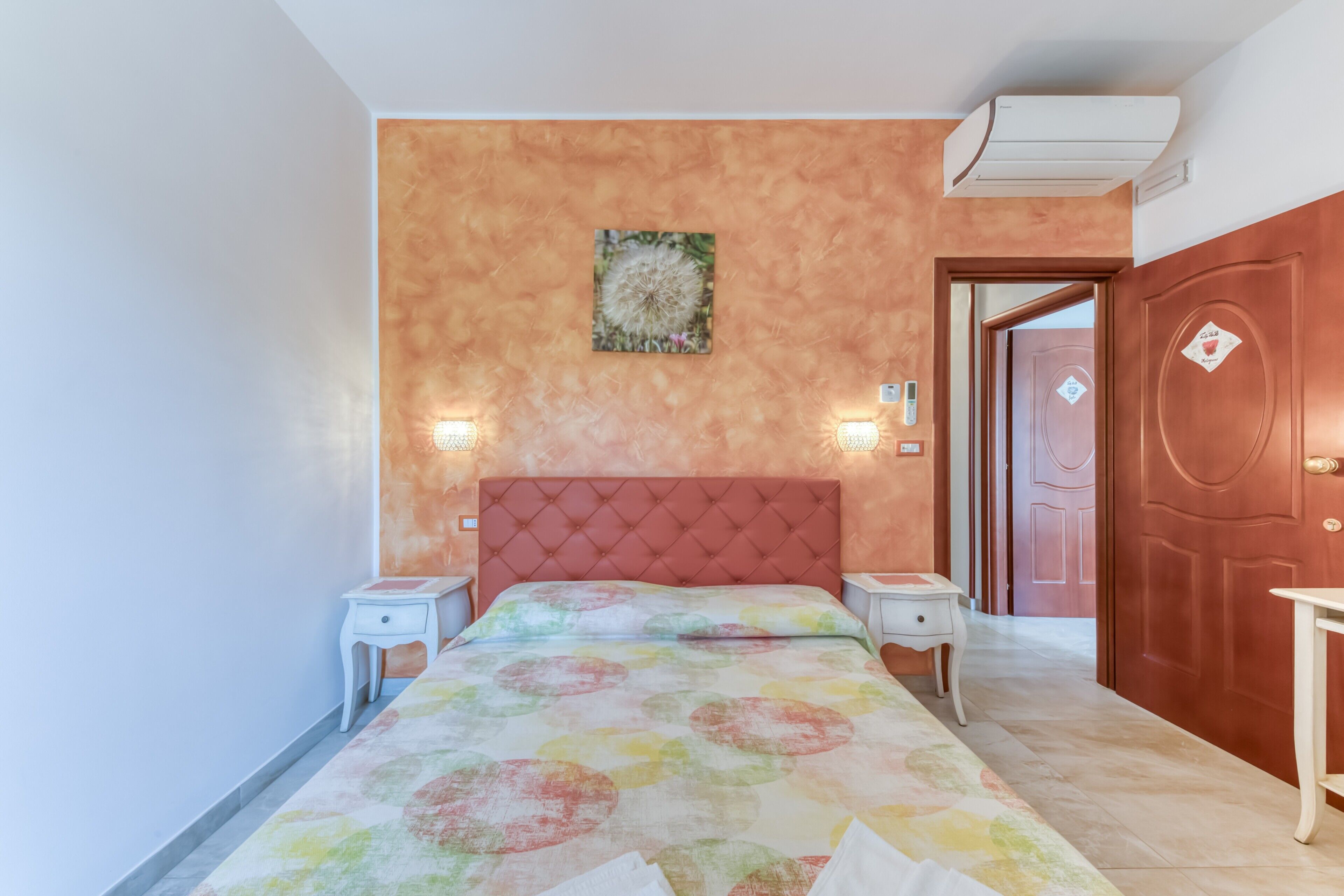 A bedroom with a tufted headboard, a bed with a colorful circular-patterned bedspread, two bedside tables, a wall-mounted air conditioner, and a door with a diamond-shaped decoration.